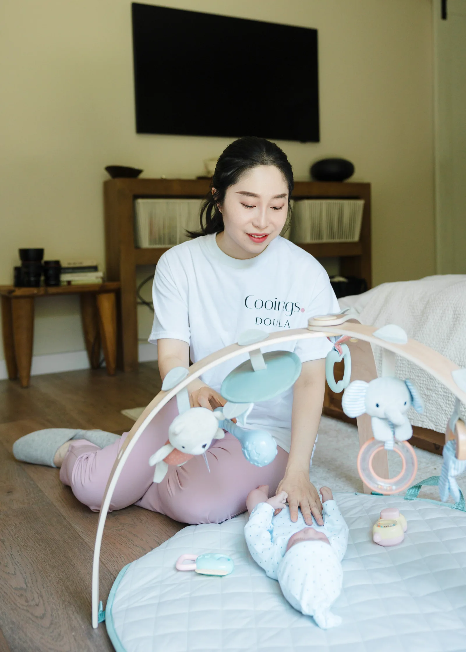 A new parent resting in a softly lit bedroom with a bowl of warm broth and a swaddled newborn nearby.