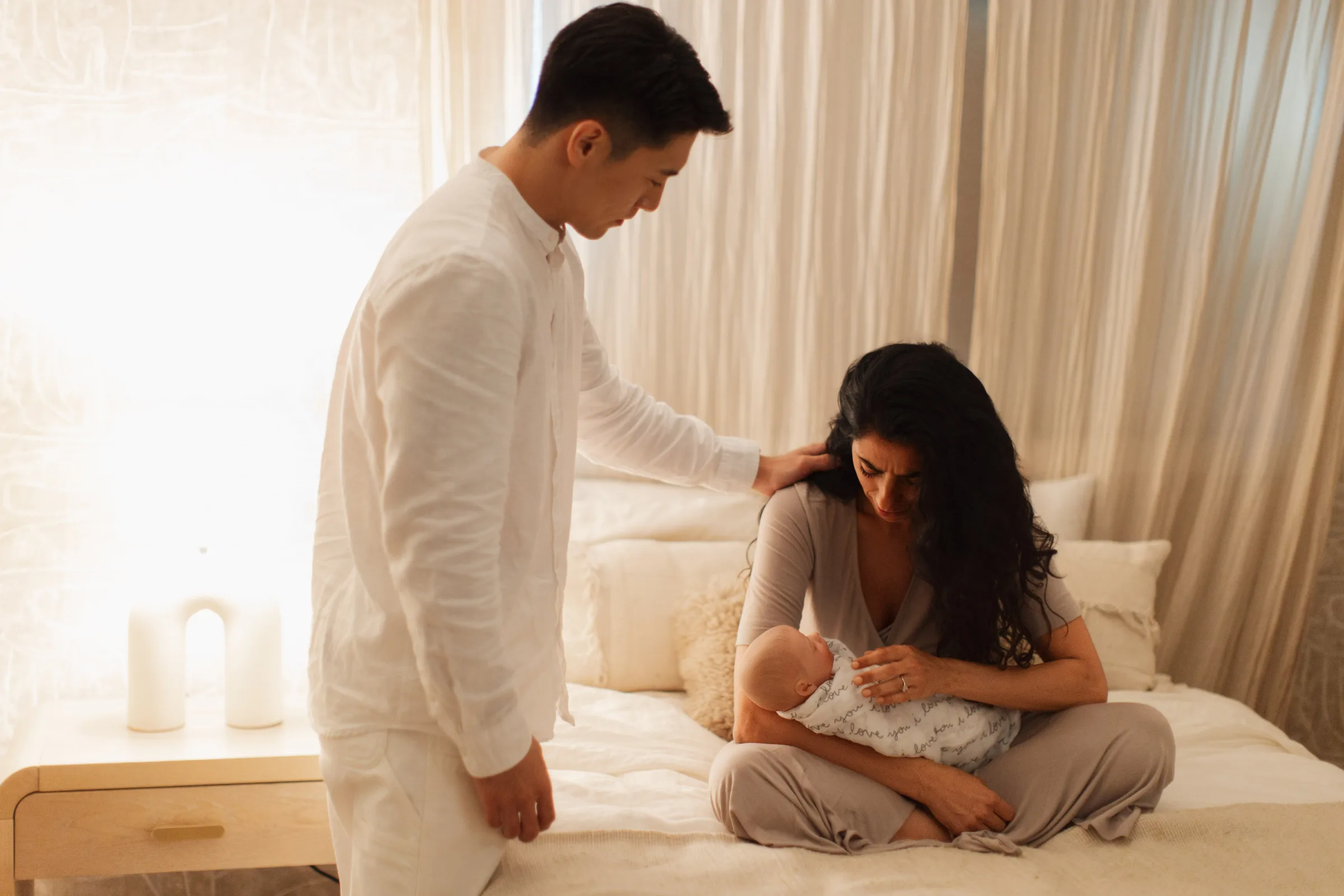 Parent holding sleeping newborn, partner standing nearby with hand on her shoulder, soft daylight through sheer curtains
